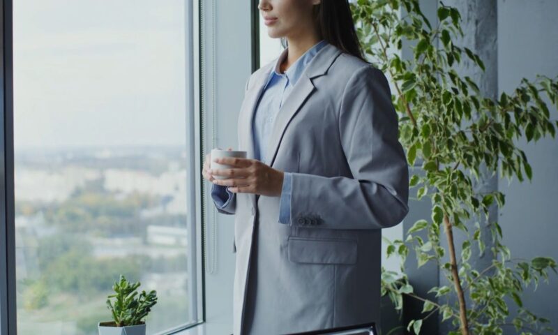 devojka stoji pored prozora u kancelariji obučena u košulju i sivi sako